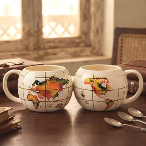 May include: Two round, cream-colored ceramic mugs featuring a colorful world map design with gold grid lines. Each mug has a curved handle and displays different continents, including Asia, Africa, and the Americas, with small text labels.