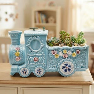 May include: A light blue ceramic train-shaped planter featuring intricate embossed lace-like patterns and small, colorful floral accents. The top of the train car is open, containing several green succulent plants nestled in soil and small decorative pebbles.