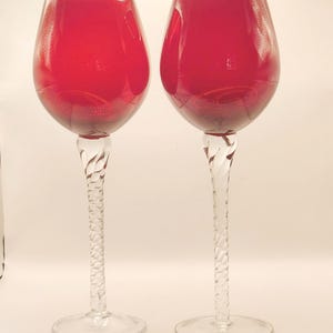 May include: Two wine glasses with deep red bowls and clear, twisted glass stems. The bowls are rounded and smooth, while the stems feature a distinct spiral pattern that connects to a circular, flat base. The glass is translucent and reflective.