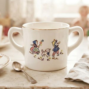 May include: A white ceramic two-handled mug featuring a gold-trimmed rim and a colorful illustration of two children and a small lamb walking through a field of yellow flowers. The mug has a smooth, cylindrical body and two curved side handles.