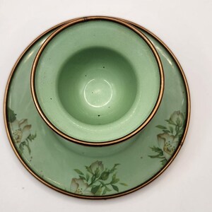 Puede incluir: Un juego de taza y platillo de t&eacute; vintage de esmalte verde. La taza se asienta sobre el platillo, ambos con un dise&ntilde;o floral y un borde color cobre. El juego es de color verde claro.
