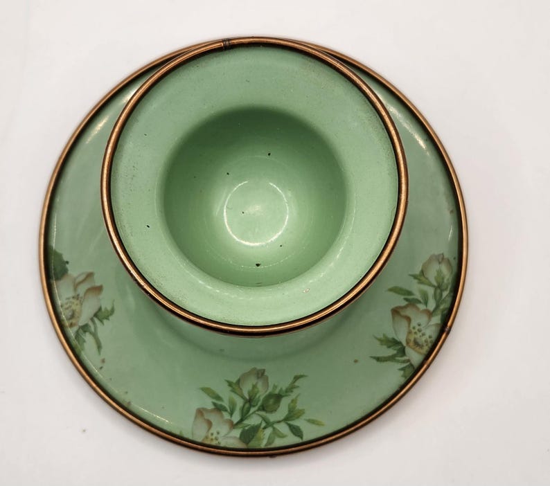 Puede incluir: Un juego de taza y platillo de t&eacute; vintage de esmalte verde. La taza se asienta sobre el platillo, ambos con un dise&ntilde;o floral y un borde color cobre. El juego es de color verde claro.