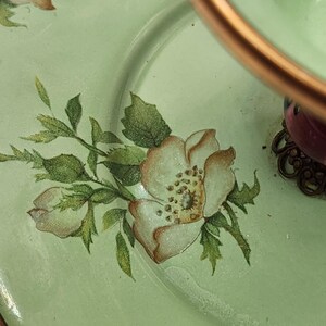 Puede incluir: Primer plano de un plato verde claro con un dise&ntilde;o floral. El plato presenta flores blancas y marrones pintadas a mano con hojas verdes. Un asa de color cobre y una base decorativa son visibles en la esquina superior derecha.