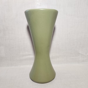May include: A sage green ceramic vase with a unique hourglass shape. The vase has a wide top and base, tapering inward at the center. The smooth, matte finish and simple design make it a versatile decorative piece.