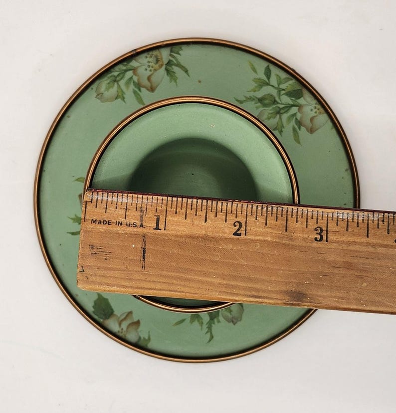 Puede incluir: Un platillo verde vintage con un dise&ntilde;o floral y ribete dorado. Una regla de madera se coloca sobre el platillo, con el texto "MADE IN U.S.A." visible. El platillo tiene aproximadamente 7.6 cm de di&aacute;metro.