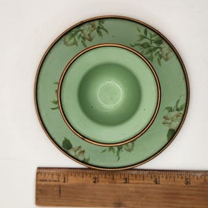 Puede incluir: Un juego vintage de huevera y platillo verdes. La huevera est&aacute; centrada en el platillo, ambos con un borde dorado. El platillo tiene un dise&ntilde;o floral con flores blancas y beige. La huevera tiene aproximadamente 5 cm de di&aacute;metro.