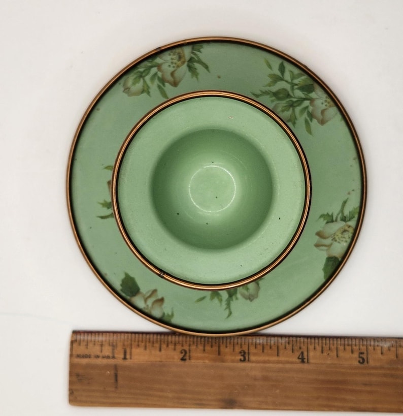 Puede incluir: Un juego vintage de huevera y platillo verdes. La huevera est&aacute; centrada en el platillo, ambos con un borde dorado. El platillo tiene un dise&ntilde;o floral con flores blancas y beige. La huevera tiene aproximadamente 5 cm de di&aacute;metro.