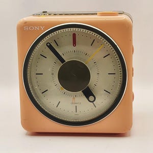 May include: A square, peach-colored Sony quartz alarm clock with a circular face. The clock features a white dial with black hour markers, thick black hour and minute hands, a thin yellow second hand, and a red alarm indicator. The word SONY is printed in white at the top left, with QUARTZ and JAPAN printed at the bottom.