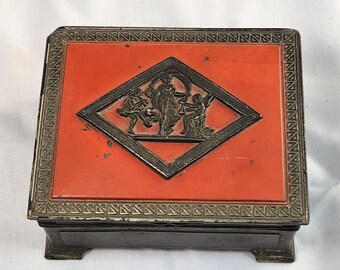 May include: A small, square, black box with a red lid. The lid has a silver border and a silver diamond-shaped design with a raised image of three figures.