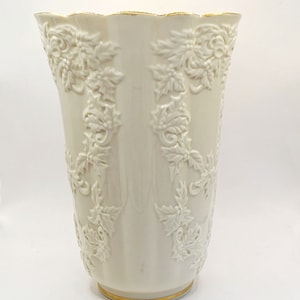 May include: A white ceramic vase with a gold rim and base. The vase is decorated with a raised design of holly leaves and berries.