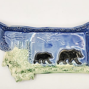 May include: A ceramic tray in the shape of the state of Montana, featuring a blue sky and green landscape. Two black bears are depicted walking across the tray. The border is decorated with paw prints.