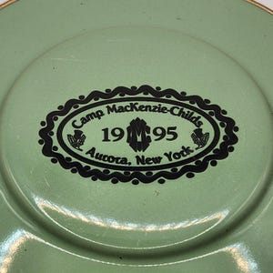 Puede incluir: Un plato verde claro con un dise&ntilde;o ovalado negro en el centro. El dise&ntilde;o incluye el texto "Camp MacKenzie-Childs 1995 Aurora, New York" en una fuente decorativa. El plato tiene un borde ligeramente elevado.