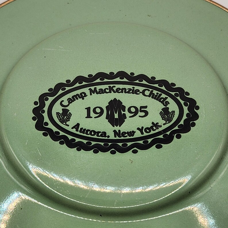 Puede incluir: Un plato verde claro con un dise&ntilde;o ovalado negro en el centro. El dise&ntilde;o incluye el texto "Camp MacKenzie-Childs 1995 Aurora, New York" en una fuente decorativa. El plato tiene un borde ligeramente elevado.
