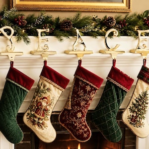 May include: Five Christmas stockings hang from gold-colored metal stocking holders that spell out the word PEACE. The stockings feature varied designs including quilted dark green velvet, cream fabric with Santa illustrations, and deep red velvet.