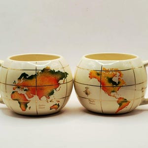 May include: Two round, cream-colored ceramic mugs featuring a world map design. The mugs are decorated with gold grid lines and colorful continents in shades of orange, yellow, and green. Each mug has a single handle and a rounded, globe-like shape.