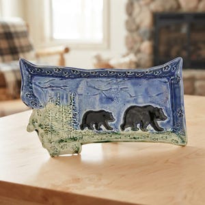 May include: A rectangular ceramic tray shaped like the state of Montana, featuring a glossy blue and green glaze. The surface displays a relief design of two black bears walking through a forest of pine trees, with paw prints along the border.