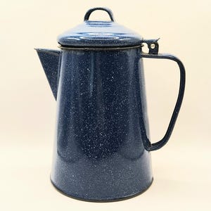 May include: A dark blue enamel coffee percolator with a white speckled pattern. The vessel features a tapered cylindrical body, a side handle, a hinged lid with a small handle, and a pointed spout. The finish is glossy and smooth throughout.