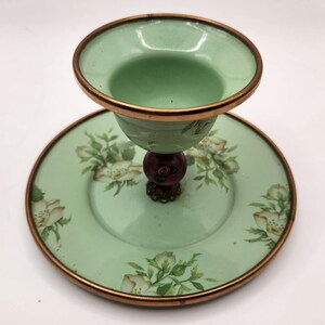 Puede incluir: Una compotera de cristal verde vintage con un platillo a juego. La compotera tiene un borde color cobre y un dise&ntilde;o floral decorativo en el platillo. El tallo presenta una cuenta roja oscura y una base ornamentada.