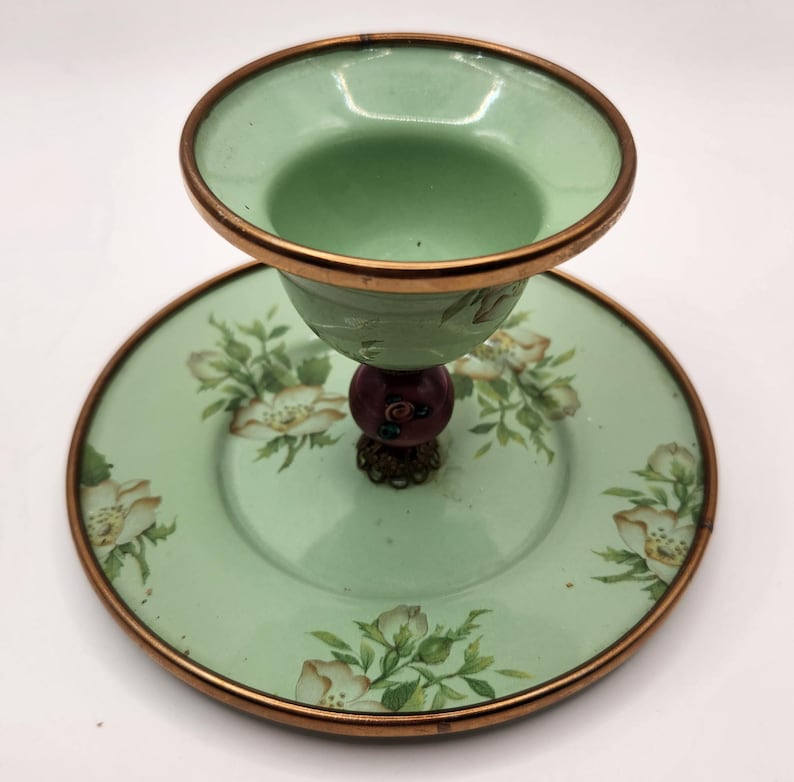 Puede incluir: Una compotera de cristal verde vintage con un platillo a juego. La compotera tiene un borde color cobre y un dise&ntilde;o floral decorativo en el platillo. El tallo presenta una cuenta roja oscura y una base ornamentada.
