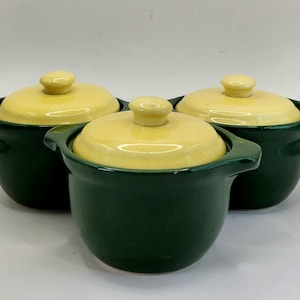 May include: Three ceramic lidded bowls. The bowls are dark green with yellow lids and small round handles. The lids have a small, round knob on top. The bowls are arranged in a group.