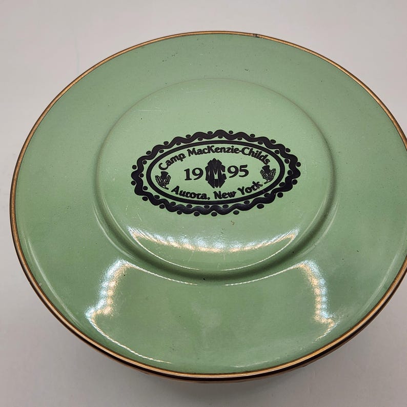 Puede incluir: Un plato redondo verde claro con un borde dorado. El centro presenta un dise&ntilde;o ovalado negro con el texto "Camp Mackenzie-Childs 1995 Aurora, New York."