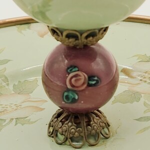 Puede incluir: Un cuenco decorativo de estilo vintage. El cuenco tiene una parte superior verde claro y una base esf&eacute;rica granate con detalles florales. La base est&aacute; soportada por una ornamentada metalister&iacute;a dorada. El cuenco se asienta sobre una bandeja con motivos florales.