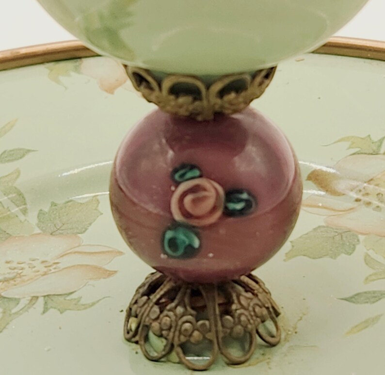Puede incluir: Un cuenco decorativo de estilo vintage. El cuenco tiene una parte superior verde claro y una base esf&eacute;rica granate con detalles florales. La base est&aacute; soportada por una ornamentada metalister&iacute;a dorada. El cuenco se asienta sobre una bandeja con motivos florales.