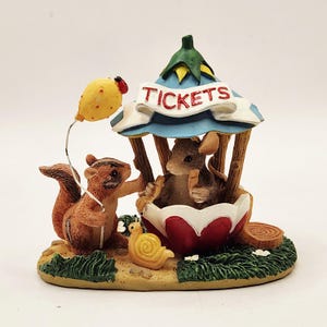May include: A decorative figurine featuring a brown squirrel holding a yellow balloon and a mouse inside a red and white ticket booth with a blue roof. The word TICKETS is printed on a white banner. A small yellow snail sits on the green base.