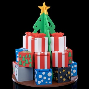 K&ouml;nnte beinhalten: Eine 3D-gedruckte Weihnachtsbaumstruktur aus gr&uuml;nem Kunststoff mit einem gelben Stern als Spitze, umgeben von einer Basis aus gestapelten quadratischen Geschenkboxen. Die Boxen haben verschiedene Farben wie Wei&szlig;, Rot, Blau und Grau mit Mustern aus Punkten, Sternen und B&auml;umen.