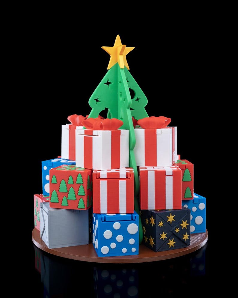 K&ouml;nnte beinhalten: Eine 3D-gedruckte Weihnachtsbaumstruktur aus gr&uuml;nem Kunststoff mit einem gelben Stern als Spitze, umgeben von einer Basis aus gestapelten quadratischen Geschenkboxen. Die Boxen haben verschiedene Farben wie Wei&szlig;, Rot, Blau und Grau mit Mustern aus Punkten, Sternen und B&auml;umen.