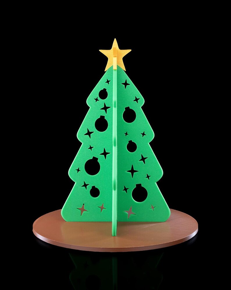 K&ouml;nnte beinhalten: Eine gr&uuml;ne, dreidimensionale Weihnachtsbaumdekoration mit ausgestanzten Stern- und Kugelformen. Der Baum ist mit einem gelben Stern gekr&ouml;nt und steht auf einem runden braunen Sockel, was ein strukturiertes und geschichtetes Erscheinungsbild ergibt.