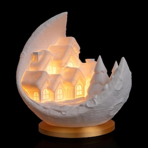 Op de afbeelding: Een witte, verlichte kerstdorpsscène in een maanvormige sculptuur. Het dorp heeft verschillende huizen met verlichte ramen en met sneeuw bedekte daken, geplaatst op een goudkleurige basis. De achtergrond is zwart.