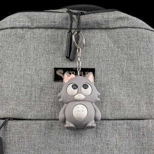 Könnte beinhalten: Ein grauer Katzen-Schlüsselanhänger mit rosa Schleife, großen Augen und weißem Bauch hängt an einer silbernen Kette. Der Schlüsselanhänger ist an einem grauen Rucksack befestigt. Der Rucksack hat ein schwarzes Etikett mit dem Wort "SOTLEL".