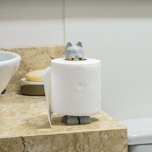 May include: A white toilet roll with a grey cat-shaped holder. The cat has yellow eyes and sits atop the roll. The holder is on a marble countertop in a bathroom setting.