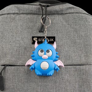 Könnte beinhalten: Ein blauer Monster-Schlüsselanhänger mit rosa Flügeln und Hörnern hängt an einem grauen Rucksack. Der Schlüsselanhänger hat einen weißen Bauch und eine kleine rosa Nase. Der Rucksack hat einen schwarzen Reißverschluss und den Text "STOLIX" auf einem schwarzen Etikett.