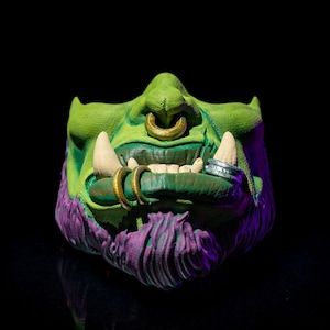May include: A green half-face mask featuring a textured purple beard, prominent white tusks, and gold-colored rings in the nose and lower lip. The mask displays detailed facial features with a dark green skin tone and a silver ring on the left tusk.