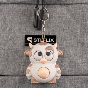 Könnte beinhalten: Ein Schlüsselanhänger mit einem Cartoon-Kuh-Design in Braun und Weiß. Die Kuh hat große Augen und kleine Hörner. Der Schlüsselanhänger ist an einem schwarzen Anhänger mit dem Text "STLFLIX" befestigt.