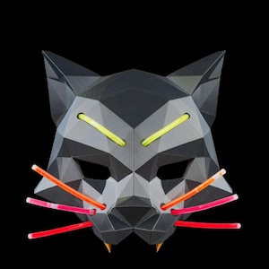 Pode incluir: Uma máscara de gato geométrica de baixo polígono em preto fosco com dois bastões luminosos amarelos brilhantes como sobrancelhas, seis bastões luminosos laranja e rosa como bigodes e dois pequenos dentes pontiagudos amarelos na borda.