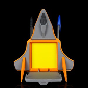 Puede incluir: Un organizador de escritorio con forma de avión de combate, que presenta un soporte para notas cuadrado de color amarillo brillante en el centro, una estructura de plástico naranja y gris, y dos ranuras verticales con un bolígrafo negro y uno azul.