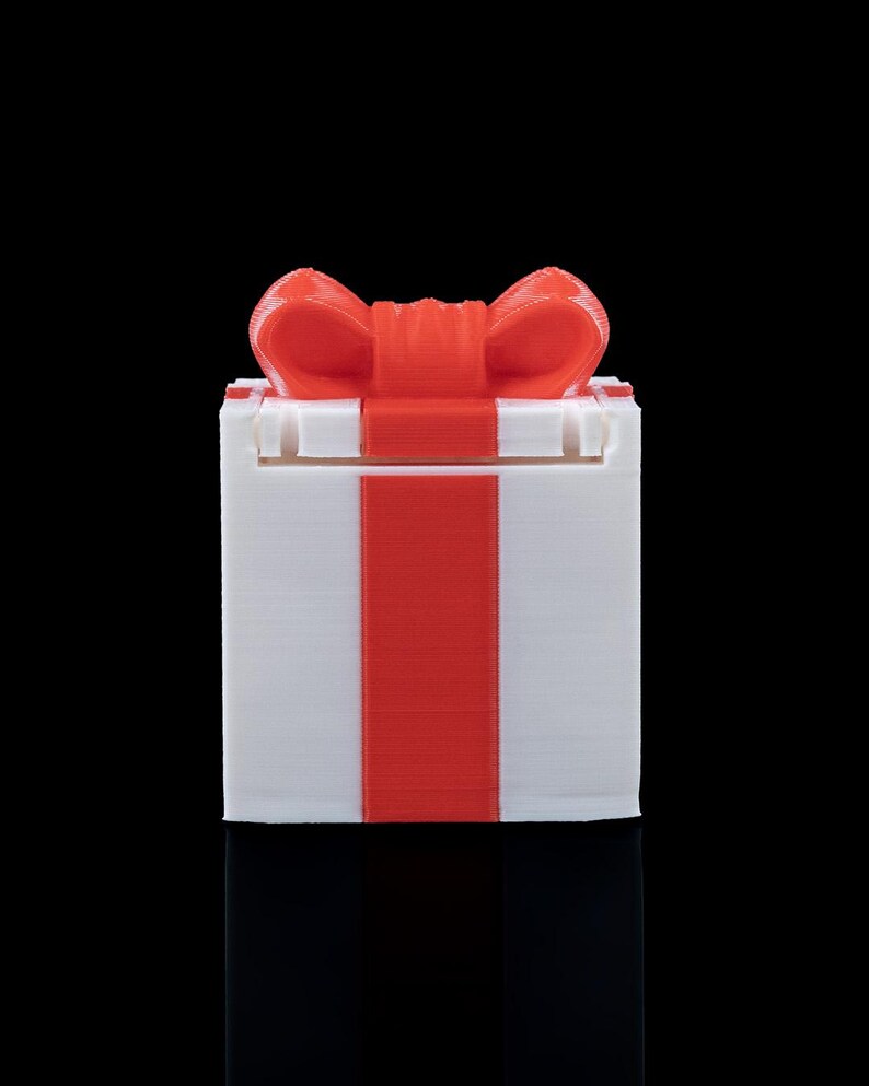 K&ouml;nnte beinhalten: Eine quadratische wei&szlig;e Box mit einem vertikalen roten Streifen in der Mitte und einer roten Schleife oben. Das Objekt weist eine sichtbare Schichtstruktur auf, die f&uuml;r den 3D-Druck typisch ist, mit klarem, geometrischem Design und flacher Basis.