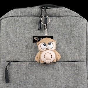 May include: A brown owl keychain with large eyes and a circular belly is attached to a gray backpack. The keychain is made of a soft material and has a metal ring. The backpack has a zipper and a small logo.