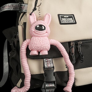 May include: A beige and black backpack with a black rectangular patch featuring the text NXN NIEXNIE. Attached to the backpack is a pink, one-eyed creature keychain with long, flexible, segmented arms and a textured, furry-looking body.