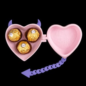 Puede incluir: Una caja rosa en forma de corazón con cuernos y cola de diablo, que contiene tres bombones envueltos en oro. La caja está abierta, revelando los bombones y el interior vacío. La cola es morada.