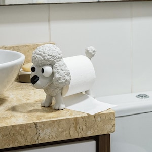 May include: A novelty white poodle-shaped toilet roll holder with a roll of white toilet paper. The poodle has a textured, curly fur design, black nose, and large, cartoonish eyes. The holder is on a marble countertop.