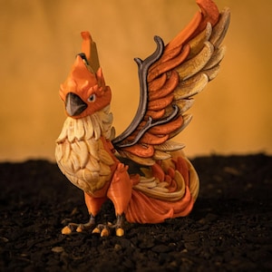 Può includere: Statuetta dettagliata di un uccello mitico, principalmente arancione con accenti di giallo, crema e nero. L'uccello ha grandi ali dettagliate e una cresta. La statuetta è su una superficie scura e strutturata.