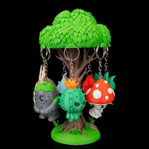 Puede incluir: Un original llavero en forma de árbol con varios llaveros temáticos de fantasía. Los llaveros incluyen una criatura de piedra gris, un cactus verde y una seta roja con lunares blancos. El árbol tiene una copa verde frondosa y un tronco marrón.