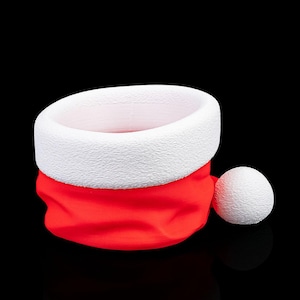 Könnte beinhalten: Ein festlicher Weihnachtsmannhut mit weißem, strukturiertem Rand und roter Krone. Ein weißer Bommel ist an der Seite befestigt. Der Hut ist vor schwarzem Hintergrund platziert.