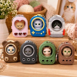 May include: Seven miniature cat keychains featuring various feline figures encased in stylised plastic housings. The designs include a pink heart-shaped case, a blue gaming console, a white rocket, and four textured cases in brown, black, green, and tan.