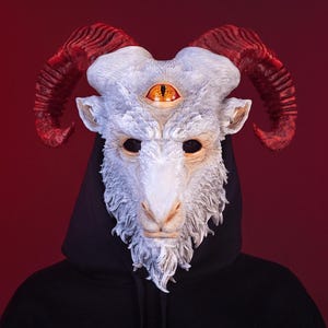 May include: A white goat mask featuring large, textured red horns and a prominent third eye centered on the forehead. The mask has detailed fur-like sculpting, dark eye sockets, and is worn over a black hooded sweatshirt against a dark red background.