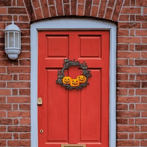 May include: A circular Halloween wreath featuring three bright orange jack-o'-lanterns with carved faces, surrounded by dark gray branches and three black cat figures with pointed ears and wide eyes, mounted on a vibrant red wooden door.